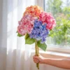 Three multicolor hydrangea stems held beside a window KAMMAK 8KPFZH002 home decor product side angle view showing silhouette